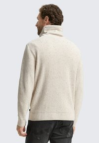 Cremefarbenes Sweatshirt mit hohem Rippenkragen, strukturiertem Strickdesign und gesprenkelten Details. Getragen mit dunklen Jeans.