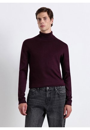 Uomo che indossa un maglione a collo alto viola scuro e jeans neri slavati, in piedi davanti a uno sfondo bianco uniforme.