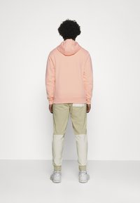 Person med lockigt hår, klädd i en ljusrosa hoodie, beiga och vita lapptäcktsbyxor och vita sneakers, står vänd mot en tom vit vägg.