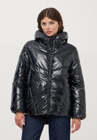 Giubbotto puffer nero con finitura lucida, cappuccio, zip verticale e motivo a rombi. Indossato sopra jeans scuri.