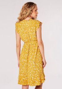 Gelbes Blumenkleid mit kurzen Rüschenärmeln, taillierter Taille, gestuftem Saum und weißen Gänseblümchenmustern, aus leichtem Stoff gefertigt.