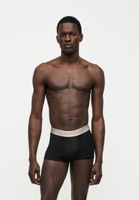Homme athlétique à la peau foncée debout, portant un boxer Calvin Klein noir, torse nu et expression faciale neutre sur fond uni.