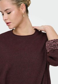 Bordowy sweter z okrągłym dekoltem i teksturowanymi rękawami, ozdobiony wielokolorowym wzorem. Tkanina wydaje się miękka z delikatnym połyskiem.