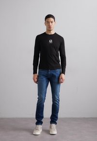 Black long-sleeve shirt with white logo on chest, paired with blue denim jeans and beige sneakers. Simple, fitted design, minimal detailing.
