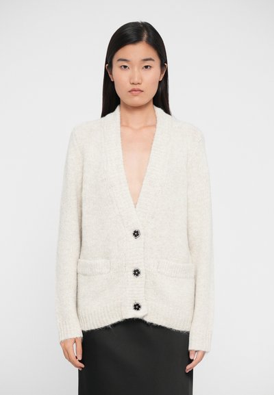Woman wearing a cream knitted cardigan with black flower-shaped buttons and two front pockets, paired with a black skirt, standing against white.