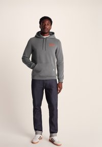 Gray hooded sweatshirt made of cotton blend material, featuring a front pocket and printed text in orange. Paired with dark denim jeans and sneakers.