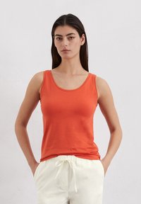 Oranges ärmelloses Tanktop aus weichem, glattem Stoff. Mit Rundhalsausschnitt und taillierter Passform, kombiniert mit hellen, lockeren Hosen.