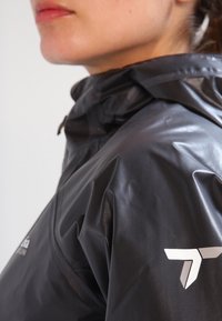 Sort vandtæt jakke med høj krave, tekstureret stof og et hvidt logo på skulderen. Nærbillede der fokuserer på materialet og designet.