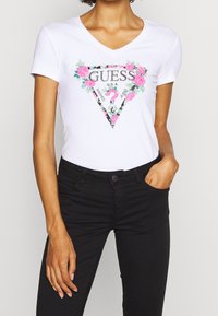 Camiseta blanca de manga corta con escote en V, que presenta un logo triangular con estampado floral y el texto "GUESS" y "WASHED JEANS."