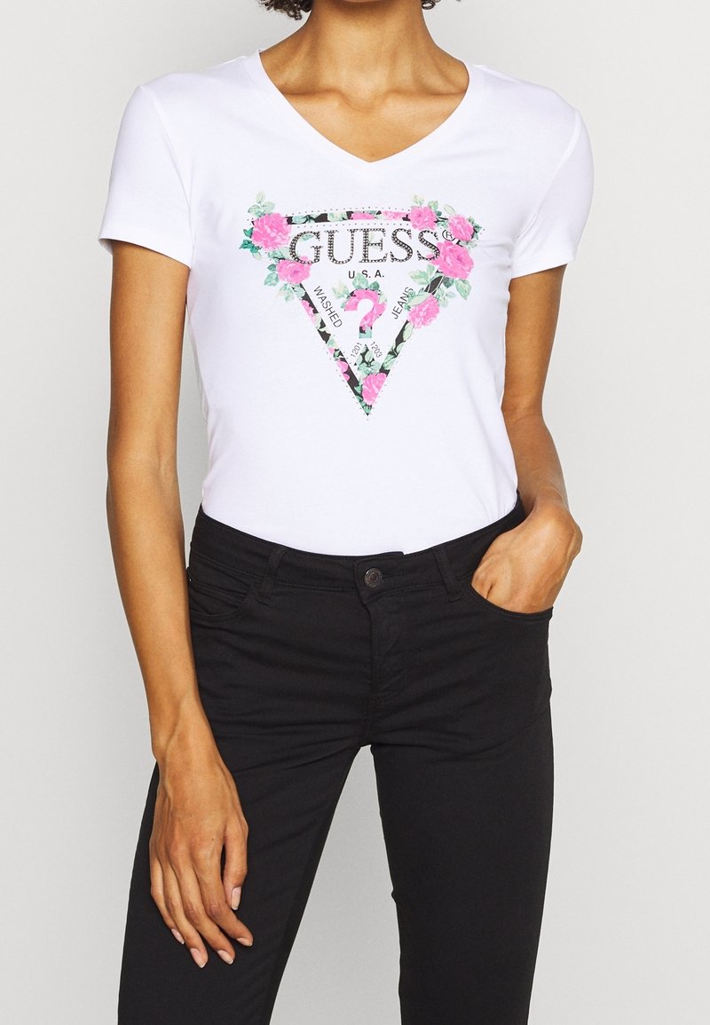 Camiseta blanca de manga corta con escote en V, que presenta un logo triangular con estampado floral y el texto "GUESS" y "WASHED JEANS."