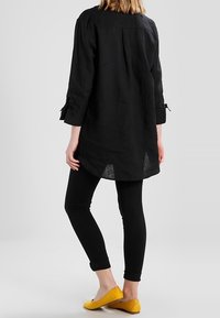 Zwarte linnen tuniek met lange mouwen, omgerolde manchetten en een iets langere achterkant. Gecombineerd met zwarte leggins en gele flats.