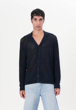 Cardigan boutonné en tricot bleu marine avec une texture en maille, des manches longues et un col classique, porté sur un jean bleu clair.