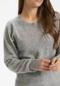 Femme portant un pull en maille gris clair avec des épaules bouffantes, des poignets et un ourlet côtelés, mains jointes près de la taille, longs cheveux bruns visibles.