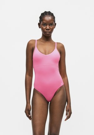 Femme aux cheveux tressés portant un maillot de bain une pièce rose à fines bretelles, debout devant un fond gris clair uni.