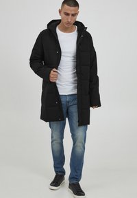 Svart polstret jakke med hette, med trykknapper, en avslappet passform og sidelommer, brukt over en hvit t-skjorte og blå jeans.
