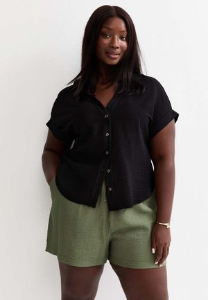 Donna che indossa una camicia nera a maniche corte con bottoni e pantaloncini verdi, in piedi con le mani in tasca davanti a uno sfondo bianco semplice.