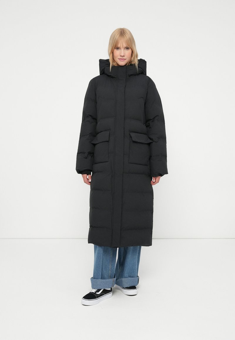 Abrigo largo acolchado negro con capucha, que presenta grandes bolsillos frontales y una cremallera delantera, llevado sobre unos vaqueros de pierna ancha y Sneakers negros.