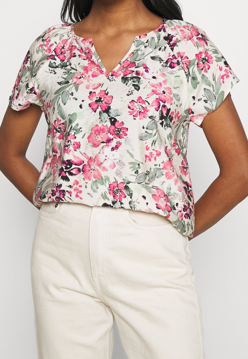 Haut à manches courtes floral avec un col en V, présentant des motifs roses et verts sur un fond beige clair. Tissu doux, coupe décontractée.