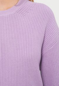 Primer plano de la parte superior del torso de una persona que lleva un suéter de punto color púrpura claro con cuello acanalado y patrón de punto texturizado.