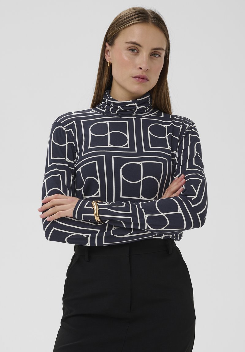 Top de cuello alto en tela azul marino con patrones geométricos blancos. Mangas largas de diseño ajustado, combinadas con una falda negra.