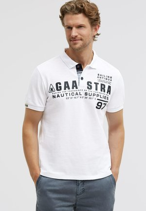 Mann mit hellem Haar und Bart, trägt ein weißes Polohemd mit schwarzem, nautisch inspiriertem Schriftzug und dunkelblaue Hose, die Hände in den Taschen.