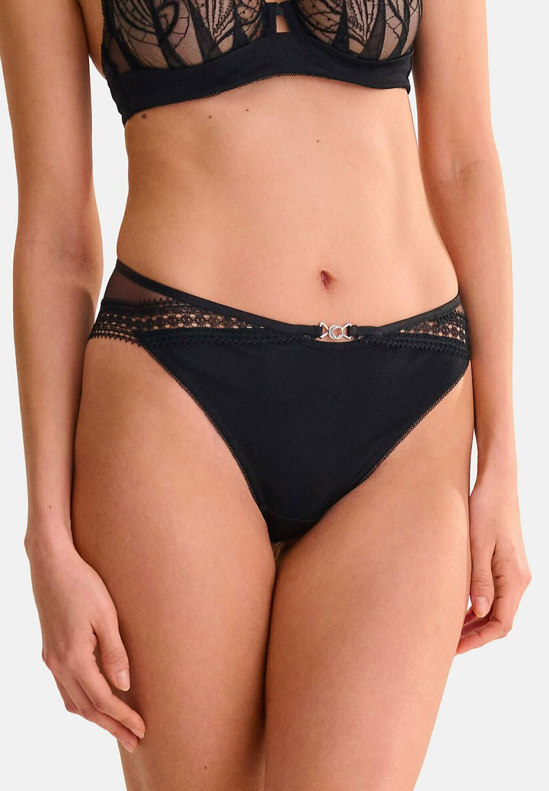 Zwarte panties van gladde stof met kantaccenten langs de tailleband en een klein zilveren detail aan de voorkant. Doorzichtige meshpanelen.