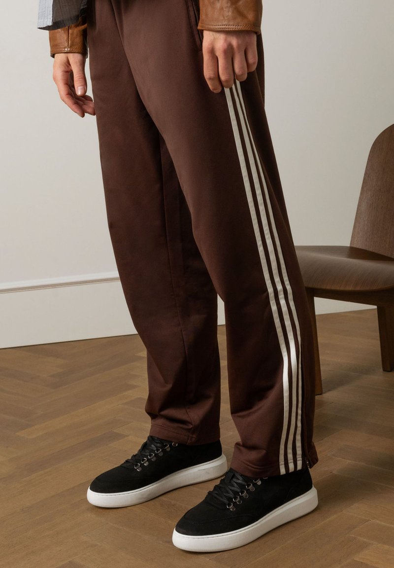 Bruine sportbroek met witte zijstrepen, gecombineerd met zwarte suède sneakers met witte zolen. Houten stoel op de achtergrond.