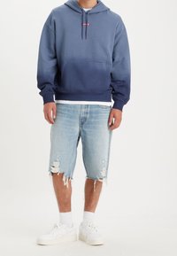Felpa navy ombre con tasca frontale e logo rosso, abbinata a pantaloncini in denim chiaro strappati e sneakers bianche.