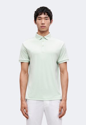 Homme portant un polo vert clair à manches courtes et un pantalon blanc, debout devant un fond uni clair, faisant face à l’avant avec une expression neutre.
