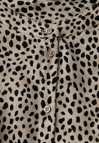 Blusa beige con stampa animale nera, caratterizzata da una lavorazione con bottoni, scollatura rotonda e tessuto liscio. I bottoni sono di colore chiaro.