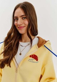 Žltá mikina na zips s hnedou kapucňou, modrými šnúrkami a vyšívaným červeným rožkom s textom "petit croissant".