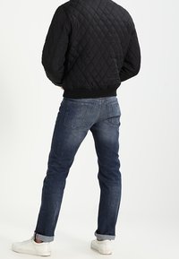 Man wearing a black quilted jacket, dark blue jeans with rolled cuffs, and white sneakers, standing facing away on a white background.