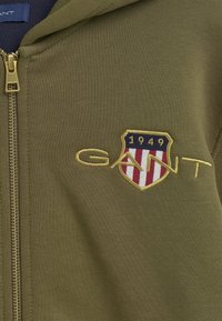 Olivově zelená zipová mikina vyrobená z hladkého materiálu, která má zlatě vyšívané logo "GANT" a štít s červenými a bílými pruhy.