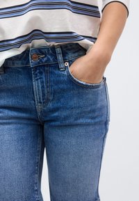 Blå denimjeans med en tætsiddende pasform, der har en kobberknap og en forlomme. Både jeansene og en hvid og blå stribet top er iført.