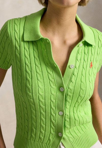 Nainen, joka pukeutunut kirkkaanvihreään, kaapelineulottuun lyhythihaiseen polopaitatakkiin, jossa on napit edessä ja pieni punainen logo rinnassa.