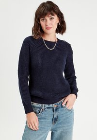 Maglione a costine navy con maniche lunghe, dotato di dettagli in metallo argentato sulla spalla e design aderente, abbinato a jeans azzurri.