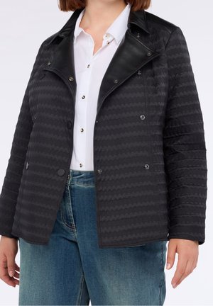 Kvinde iført en sort quiltet jakke med læderkrave over en hvid skjorte med knapper og blå jeans, stående mod en ensfarvet baggrund.