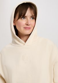 Kremowa bluza z kapturem, wykonana z miękkiego materiału, z delikatnie haftowanym logo na piersi oraz regulowanym kapturem.