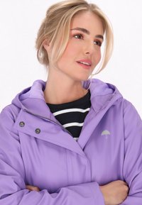 Violetinė vandeniui atspari striukė su kapišonu, puošta snapelių segtukais ir mažu lietaus skėčio logotipu. Dėvima ant tamsiai mėlynos juostuotos marškinių. Lygus audinys.