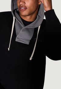 Joven vestido con una sudadera negra con capucha gris, ajustándose la capucha con una mano, labios ligeramente entreabiertos.