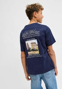 Tamsiai mėlynas medvilnės marškinėliai, kurių nugarinėje pusėje yra grafikos dizainas. Baltas tekstas ir fotopaveikslas su miesto panorama centre.