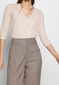 Ljus beige stickad cardigan med V-ringning och trekvartsärmar, utrustad med ett zickzackmönster. I kombination med höga rutiga byxor.