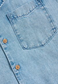 Lichtblauwe denim overhemden met een patchzak en twee houten knopen. De stof heeft een lichte textuur met vervaagde gebieden.