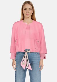 Veste rose en suédine synthétique avec un col rond, deux poches avant et des manches cloche trois-quarts. Une écharpe à motifs souligne la taille.