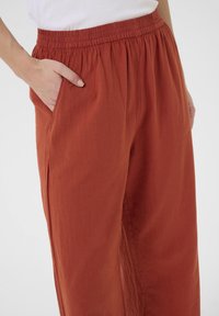 Pantalon en tissu léger couleur rouille avec une taille élastique et des poches latérales, présentant une coupe ample et une texture douce.