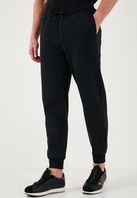 Schwarze Jogginghose mit einer Kordelzugtaillierung, seitlichen Taschen, schmal zulaufenden Beinen und gerippten Bündchen, kombiniert mit schwarzen und braunen Sneakers.