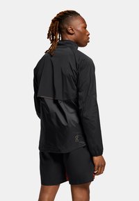 Schwarze leichte Jacke mit einem Klappen-Design auf der Rückseite, mit glatter Textur und subtiler Markenkennzeichnung. Kombiniert mit schwarzen Shorts.