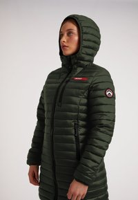 Zelená péřová bunda s kapucí, která má přední zip, panelové švy, boční kapsy a červenou nášivku na hrudi.