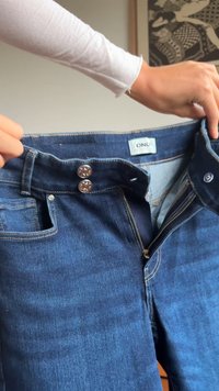 Mãos segurando um par de jeans azuis abertos, com o fecho da bragueta aberto e dois botões fechados, mostrando a etiqueta da marca "ONLY" no interior.