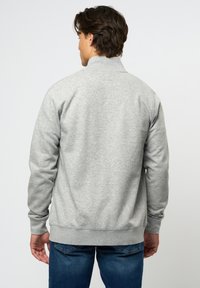 Grå hettegenser med høy krage, ribbede mansjetter og nederkant. Stoffet ser mykt ut med en glat overflate. Brukt med blå jeans, som viser en uformell stil.